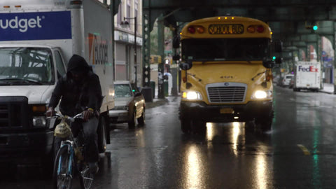 man riding bicycle with yellow school bus driving in rain under bridge in Brooklyn in slow motion