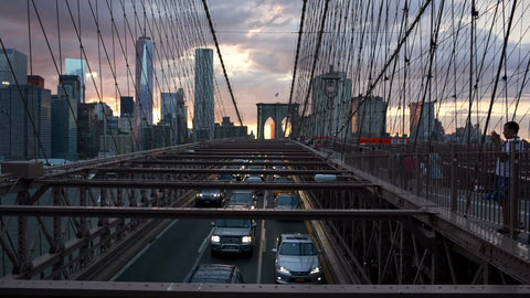 cars driving slowly in traffic over Brooklyn Bridge at sunset with Manhattan skyline in background - Freedom Tower and skyscrapers in New York City