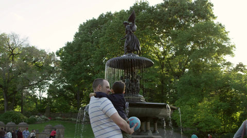 father holding son in Central Park looking at Bethesda angel statue with wings and fountain with water in summer