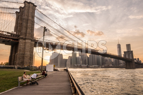 people on benches in Brooklyn Bridge Park enjoying summer view of Manhattan skyline and East River water at sunset in NYC