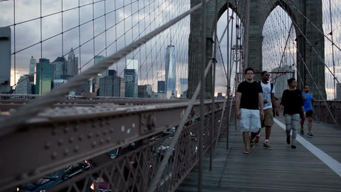 people walking across Brooklyn Bridge with Manhattan skyline and Freedom Tower in distance at sunset in summer