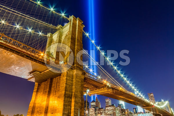 Brooklyn Bridge at night with 911 beams over Manhattan skyline, upward angle under beautiful evening sky with lights