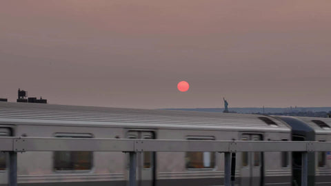 elevated subway train in Brooklyn at sunset with orange sun in sky and Statue of Liberty in distance