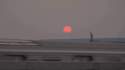 subway train passing on elevated track with orange sun and Statue of Liberty in background at sunset in Brooklyn
