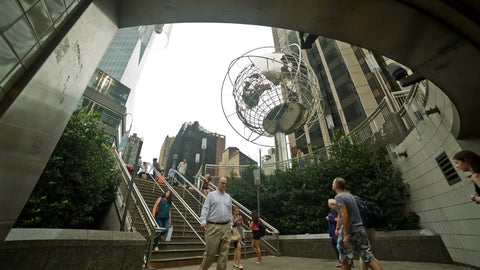 people entering and exiting Columbus Circle subway station stairs with Steel Globe Sculpture overhead on summer day in NYC