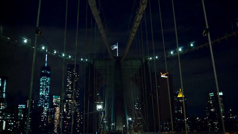 American flag atop Brooklyn Bridge with Freedom Tower and Manhattan skyline skyscrapers in background at night in NYC
