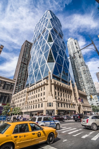 taxi cab and NYPD police vehicle driving past Hearst Tower - skyscraper in Midtown Manhattan - odd building over blue sky