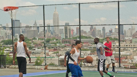 kids playing basketball on summer day with view of Manhattan skyline with Empire State Building in NYC through fence