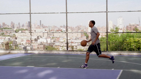 kid dribbling toward basket and shooting and scoring layup on basketball courts outside with Manhattan skyline in background through fence