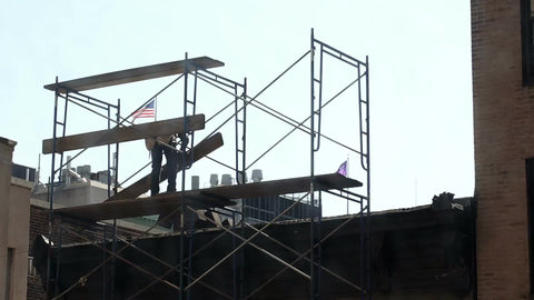 construction worker laying wood plank on scaffolding at top of building, man in hardhat on rooftop with American flag