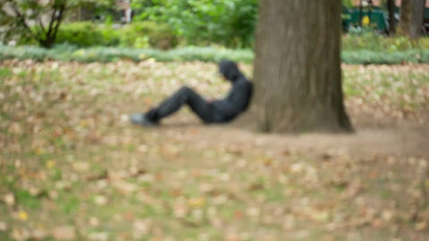 homeless man sitting under tree in black hoodie in Washington Square Park in fall - NYC