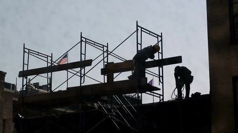 silhouette of construction workers in hardhats on scaffolding at top of building with American flag waving in NYC