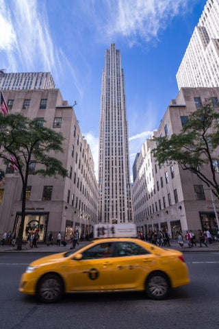 taxicab driving past Rockefeller Center on beautiful bright sunny day with blue sky and clouds in Midtown Manhattan