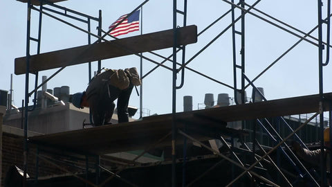 construction worker in hardhat hammering scaffolding on rooftop of building with American flag waving in NYC