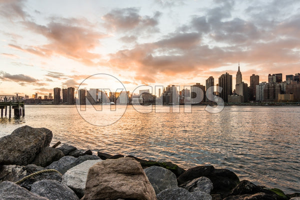 Manhattan skyline viewed from Brooklyn with rocks in foreground at sunset - beautiful orange sky and clouds over East River water