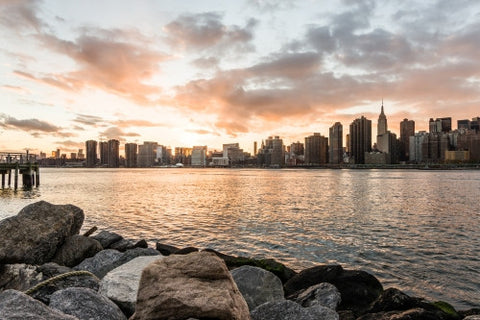 Manhattan skyline viewed from Brooklyn with rocks in foreground at sunset - beautiful orange sky and clouds over East River water