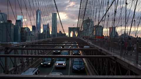 cars driving in slow traffic across Brooklyn Bridge at sunset with Manhattan skyline in background in NYC