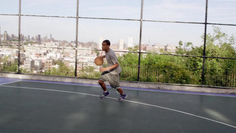 kid dribbling to basket and scoring reverse layup with left hand on basketball court with Manhattan skyline view in summer