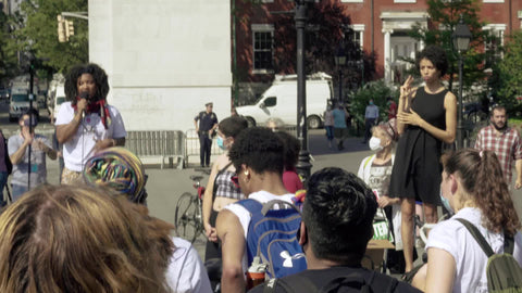 Black leader speaking about protest before crowd with sign language translator signing in Washington Square Park