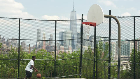 little kid practicing basketball on hot summer day with view of Freedom Tower in NYC through fence